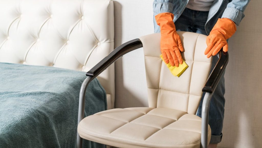 A person wearing orange gloves cleaning a beige leather chair with a yellow cloth in a modern bedroom.