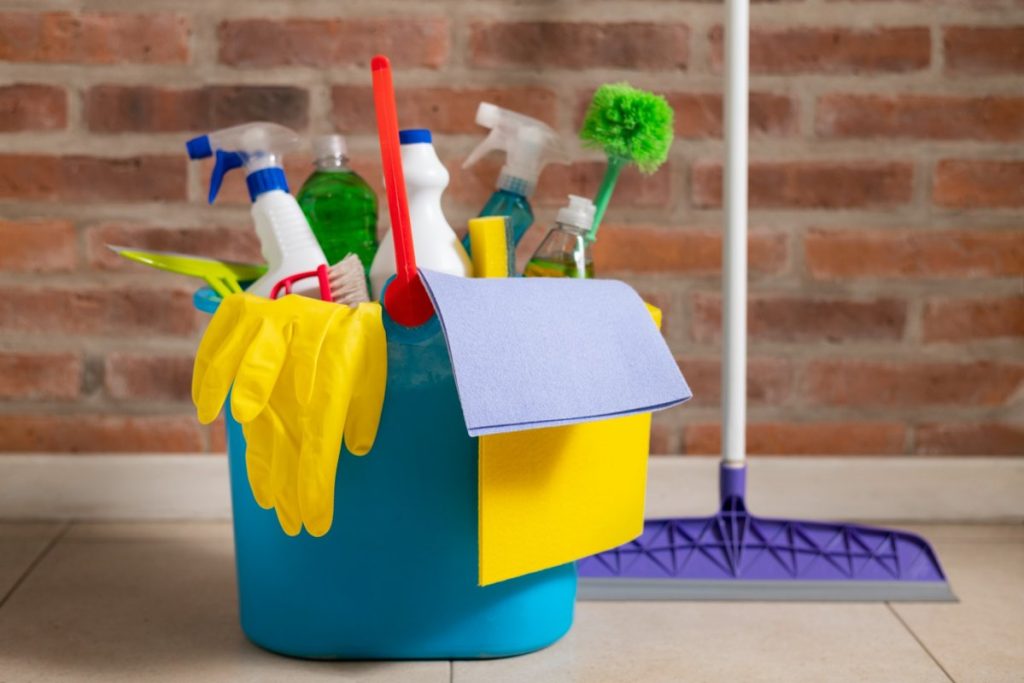 A bucket filled with essential cleaning tools including gloves, sprays, and cloths in front of a brick wall.